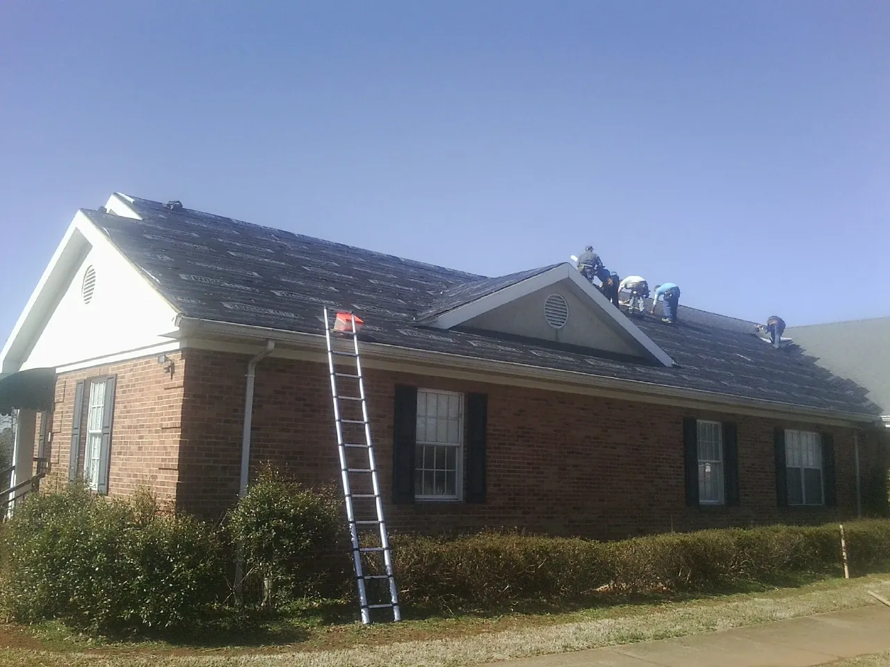 Crew tearing shingles off a brick ranch roof with a ladder set — Emergency Roof Repair work by our crew in Port Huron