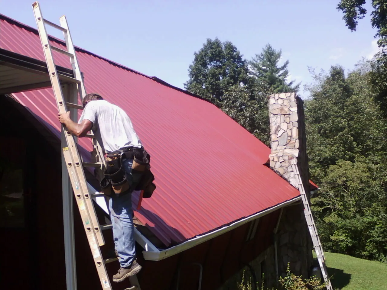 Roofer on a ladder beside a red metal roof and stone chimney — Soffit & Fascia Repair by Roofing Brothers in Port Huron